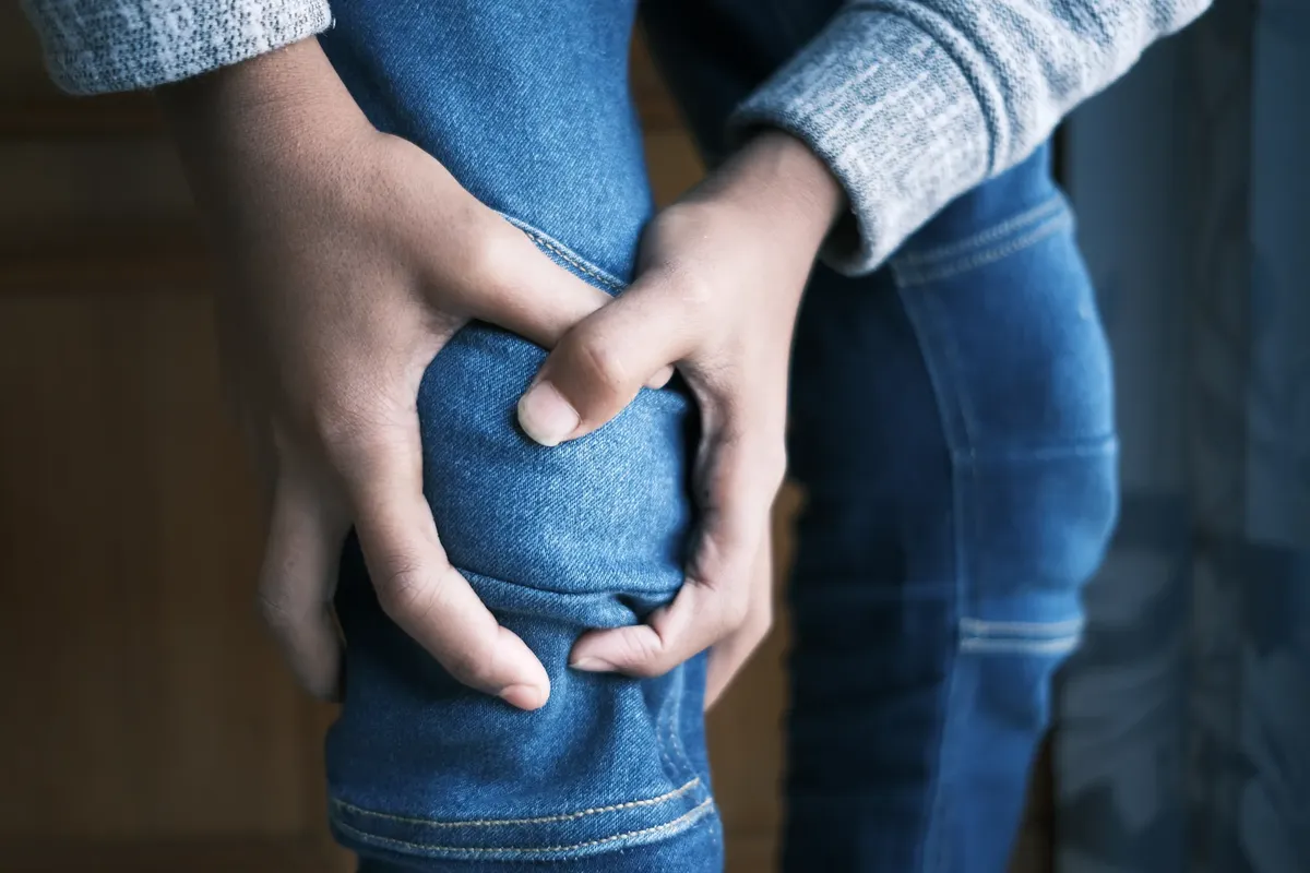 Close-up image of hands holding a knee in jeans, indicating pain or injury.